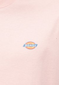 Ανοιχτό ροζ βαμβακερό μπλουζάκι με κυκλικό μπλε και πορτοκαλί λογότυπο "Dickies" στην αριστερή πλευρά του στήθους. Λείο υλικό, στρογγυλή λαιμόκοψη.