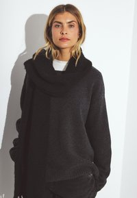 Czarny, oversize sweter z teksturowanym wykończeniem i dużym kominem. Model nosi go na białej bluzce. Miękka tkanina, prążkowane mankiety.