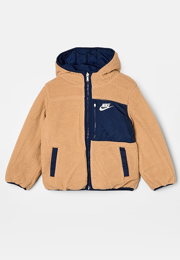 Veste en fausse fourrure beige avec une capuche et des panneaux bleu marine, fermeture éclair sur le devant, poches latérales et logo Nike blanc sur la poitrine.
