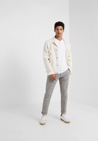 Casaco de ganga bege claro sobre uma camisa henley branca, combinado com calças cinzentas aos quadrados e ténis brancos com detalhes castanhos.