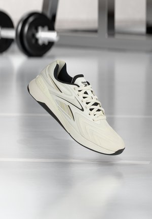 Biało-czarny but do biegania Reebok unoszący się w pomieszczeniu na błyszczącym podłożu, w tle rozmazane ciężarki siłowni.