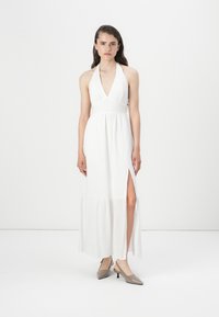 Hvit halterneck maxi-kjole med dyp V-hals, sideslitt og lagdelt kant. Modellen har på seg grå hælede mules. Enkel, flytende design.