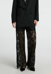 Mustat pitsihousut, joissa on leveä lahjasuunnittelu, monimutkaiset kukkakuviot ja läpikuultava rakenne, yhdistettynä ryhdikkääseen mustaan bleiseriin.