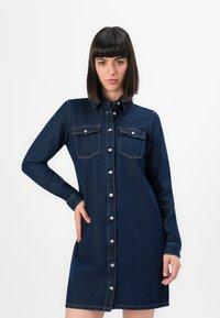 Femme avec des cheveux noirs raides portant une robe en denim bleu foncé à boutons, à manches longues et avec des poches sur la poitrine, posant avec une main sur la hanche.