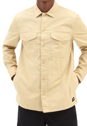 Homme portant une chemise beige à boutons avec deux poches à rabat sur la poitrine et les mains dans les poches latérales, assortie à un pantalon noir.