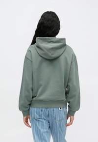 Groene hoodie met een ontspannen pasvorm, ribbelrand en manchetten. De achterkant heeft een comfortabele capuchon, gecombineerd met lichtblauwe gestreepte jeans.