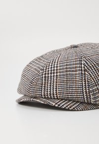 Flat cap made of textured fabric in a brown, white, and orange check pattern. Features a rounded top and a small, stiff brim.