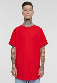 Camiseta de algodón roja con diseño oversize, mangas cortas y cuello redondo. Presenta tatuajes visibles en los brazos.
