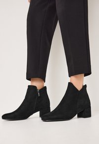 Black suede ankle boots with a pointed toe, low block heel, and side zipper. Smooth texture with a modern, minimalist design.