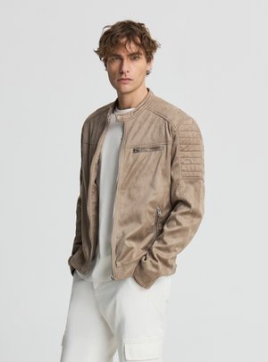 Giovane uomo con capelli ricci che indossa una giacca marrone chiaro con zip sopra una maglietta bianca e pantaloni bianchi, in posa con le mani nelle tasche.