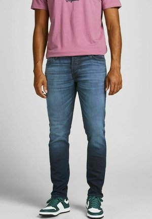 Mężczyzna w wyblakłych niebieskich dżinsach, zielono-białych sneakersach i różowej koszuli stojący na jednolitym tle.