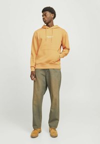 Oranje hoodie met een voorzak en wit logo, gecombineerd met loszittende, vervaagde spijkerbroeken en tan laarzen. Soepele stoftextuur.