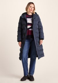 Woman wearing a long navy quilted coat over a multicolored sweater, blue jeans, and black ankle boots, standing against a neutral background.