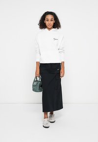 Hanorac alb cu glugă cu textul "Regular", fustă maxi neagră cu buzunar lateral, geantă verde și adidași gri. Design simplu, stil casual.