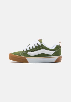 Zapatillas de ante verde con un diseño de olas blancas, cordones blancos y suela de goma marrón. Costuras destacadas y una pequeña lengüeta en el talón.
