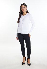 Cardigan bianco a maniche lunghe con chiusura a bottoni e polsini a costine, abbinato a leggings neri e scarpe con tacchi alti nere. Tessuto liscio.
