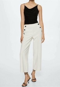 Femme portant un haut drapé sans manches noir, un pantalon blanc à taille haute et jambes larges avec des boutons noirs, et des sandales à talons hauts noires à lanières.