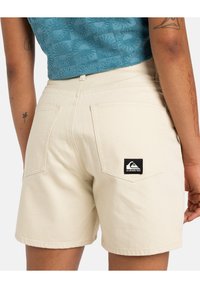 Bežid denim lühikesed püksid kahe tagataskuga, millel on must logo silti. Materjal tundub tugev ja tekstureeritud viimistlusega.