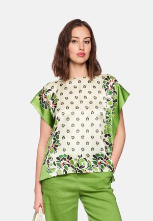 Woman wearing a green and white floral patterned blouse with matching green pants, standing with one hand in pocket and holding a white bag.