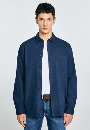 Granatowa koszula na guziki z miękkiego materiału, z kieszenią, kołnierzykiem i długimi rękawami, noszona na białej koszulce i dżinsach.