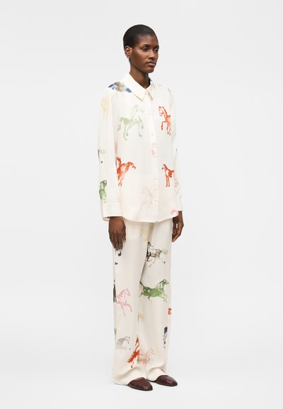 Nainen, joka pitää päällään valkoista löysää paitaa ja housuja, joissa on monivärisiä hevoshahmokuvia, yhdistettynä tummiin teräviin kenkiin, seisoo valkoisella taustalla.