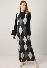 Nő áll, fekete hosszú ujjú inget visel, amely alatt ujjatlan, argyle mintás ruha van, krémszínű bokacsizmában, oldalra néz.