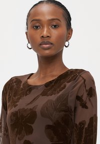 Καφέ τοπ με λουλουδάτο μοτίβο και απαλή υφή. Διαθέτει διαφανή μανίκια, στρογγυλή λαιμόκοψη και διακριτική λάμψη. Ασημένια σκουλαρίκια στεφάνια συμπληρώνουν την εμφάνιση.