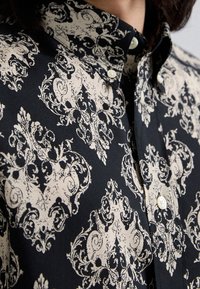 Camicia button-up nera con motivi ornamentali crema. Presenta un colletto classico e bottoni bianchi. Il tessuto appare liscio e leggero.