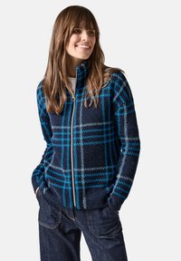 Fleece takki, jossa on vetoketju, tummansininen ja sininen ruutukuosi, pehmeä pinta ja korkea kaulus. Päällä valkoinen paita ja tummansiniset farkut.