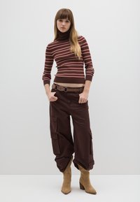 Braun gestreifter Rollkragenpullover mit langen Ärmeln, kombiniert mit locker sitzenden braunen Cargohosen mit Seitentaschen und einem braunen Gürtel. Braune Absatzstiefel.