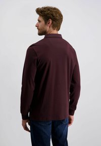 Mann mit kurzen braunen Haaren und Bart, der ein langärmliges Shirt in dunklem Burgunder und eine blaue Jeans trägt, mit dem Rücken zu einer schlichten Hintergrund.