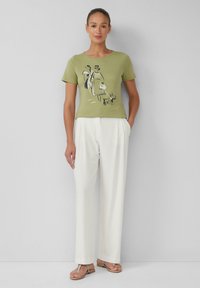 Olijfgroene t-shirt met korte mouwen en een zwart-witte grafische afbeelding van figuren, gecombineerd met witte broek en bruine sandalen.