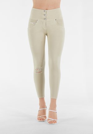Leggings a vita alta di un leggero colore beige, con chiusura a tre bottoni, tasche frontali e un ginocchio rovinato. Texture liscia con leggera elasticità.