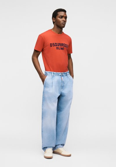 Άνδρας που φοράει πορτοκαλί μπλουζάκι DSQUARED2 Milano, ανοιχτό μπλε τζιν με φαρδιά μπατζάκια και κρεμ αθλητικά παπούτσια, στέκεται με τα χέρια στις τσέπες σε άσπρο φόντο.