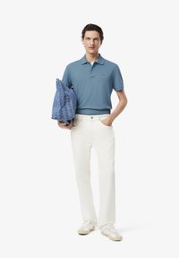 Jasnoniebieska koszulka polo z małym logo, białe spodnie szerokie oraz kremowe trampki. W lewej ręce trzymana jest wzorzysta niebieska torba.