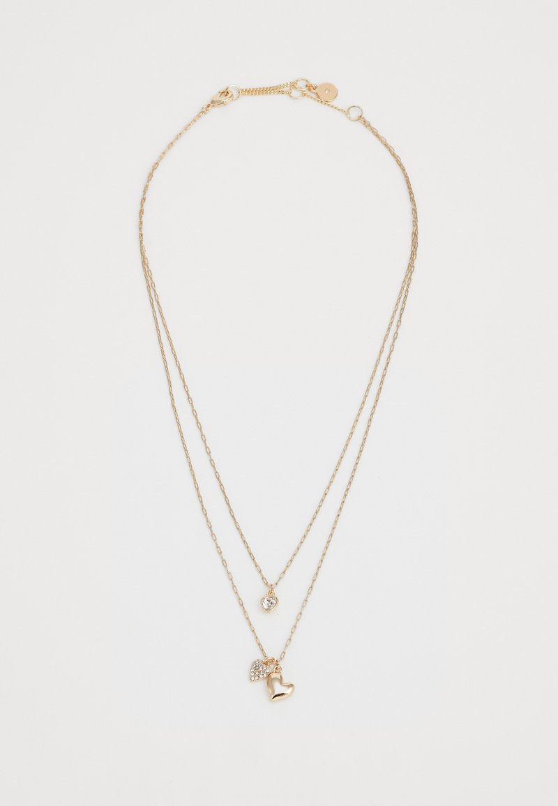 Collier en métal doré à plusieurs couches avec deux chaînes. Il présente un petit pendentif en forme de cœur en cristal et un charm en forme de cœur poli. Longueur ajustable.