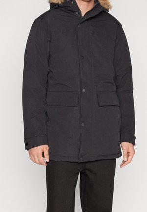 Man draagt een zwarte winterparka met een capuchon met bontvoering, voorsnapknopen, klepzakken en zwarte broek tegen een effen achtergrond.