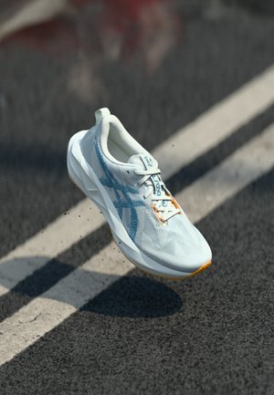 Baskets de sport blanches avec un dessus texturé présentant des accents bleu clair, une semelle orange et une languette de renforcement au niveau du talon ; lacets illustrés.