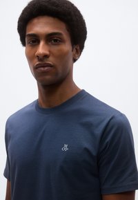 Granatowy bawełniany t-shirt z okrągłym dekoltem, krótkimi rękawami i małym białym logo "M OP" wyszytym na lewej piersi. Gładka tekstura.