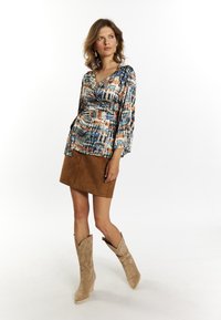 Top avvolgente multicolore con maniche lunghe, abbinato a una minigonna in suede marrone e stivali da cowboy beige con dettagli ricamati.