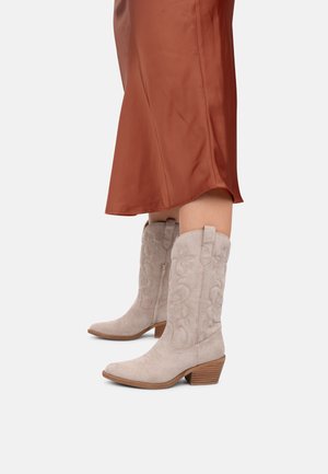 Botas de cowboy de ante beige con bordado floral, puntera afilada y tacón de madera apilada, combinadas con una falda fluida de color óxido.