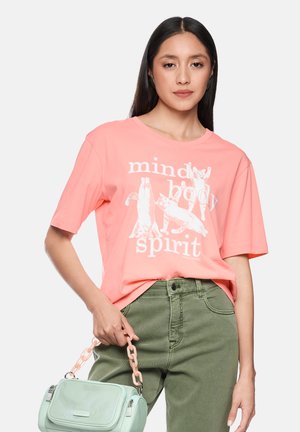 Frau trägt ein korallenrotes T-Shirt mit dem Schriftzug "mind body spirit" und Katzenillustrationen, gesteckt in olivgrüne Hosen, hält eine mintgrüne Handtasche.