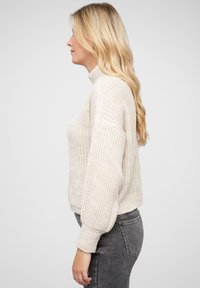 Light beige knitted sweater with a high neck, drop shoulder design, and ribbed cuffs. Worn with dark gray fitted jeans.