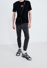 Černé tričko s malou grafikou, šedé tepláky, černá crossbody taška a černé/šedé tenisky s žlutými detaily.