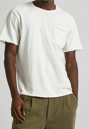 T-shirt en coton blanc à manches courtes, col rond et une seule poche poitrine. Porté avec un pantalon texturé vert olive.