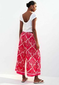 Leichtes wei�ßes Oberteil mit Rüschensäumen und offenem Rücken, kombiniert mit weit geschnittenen, rosa floral gemusterten Hosen. Flache Sandalen runden das Outfit ab.