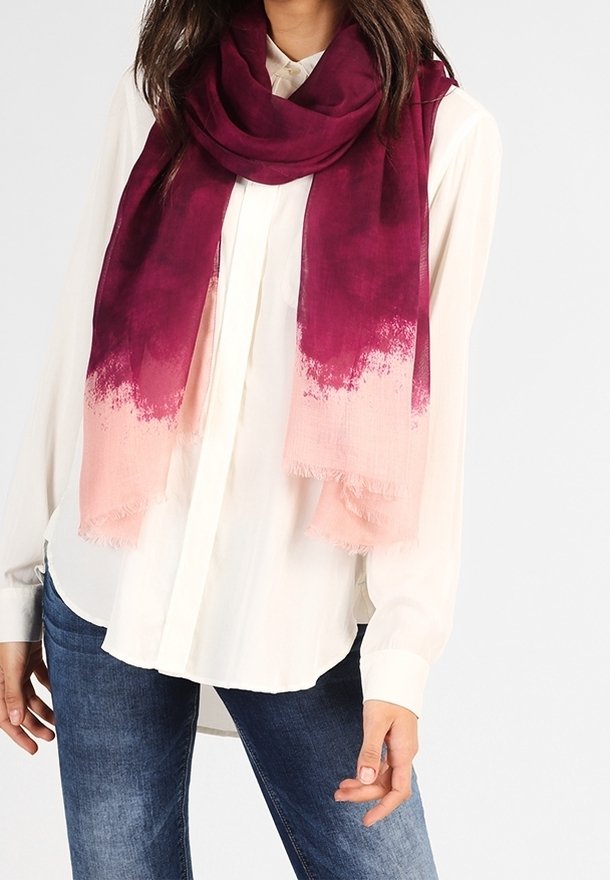 Femme portant une chemise blanche boutonnée, un jean bleu et une grande écharpe dégradée allant du rouge foncé au rose clair autour du cou.