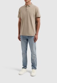 Beige poloshirt met een kraag, een knoopsluiting met drie knopen en een subtiele merklogo. Gecombineerd met lichtblauwe jeans en witte sneakers.
