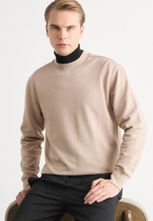 Beige crewneck sweater with long sleeves and ribbed collar, paired with a black turtleneck. Smooth texture, minimal design, and fitted shape.