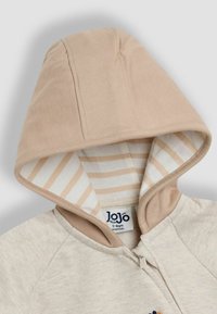 Haut à capuche beige zippé avec une capuche marron clair, présentant une doublure rayée. Tissu en coton doux avec des détails brodés sur le devant.